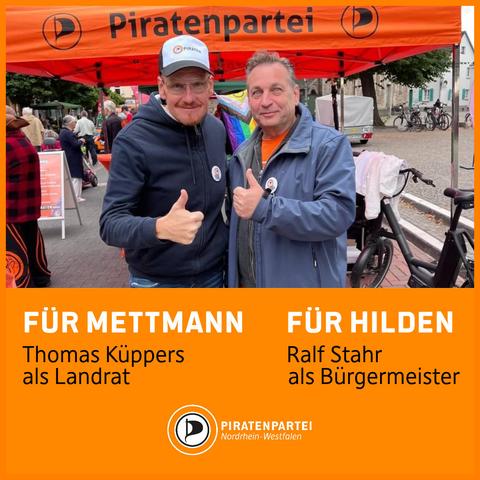 Bildtext: Für Mettmann Thomas Küppers als Landrat. Für Hilden Ralf Stahr als Bürgermeister. Piratenpartei Nordrhein-Westfalen.
Bild: Foto von Thomas Küppers und Ralf Stahr vor einem Piratenpartei-Infostand. Beide halten lächelnd ihre Daumen hoch.