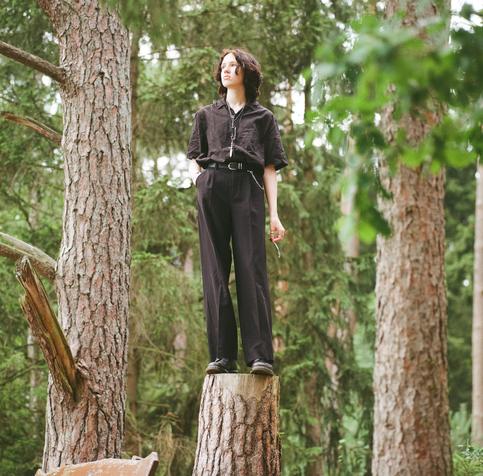 Sky steht im Wald auf einem abgesägten Baumstumpf, eine Feder in der Hand, schwarze Klamotten