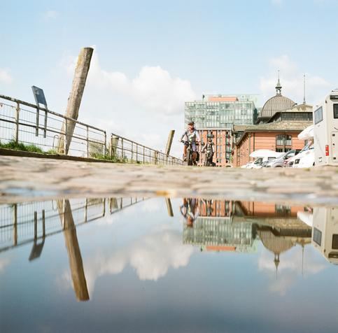 Fischmarkt, Adobe Office Hamburg in der Ferne, im Vordergrund eine Pfütze mit Reflektion, zwei Radfahrer kommen der Kamera entgegen