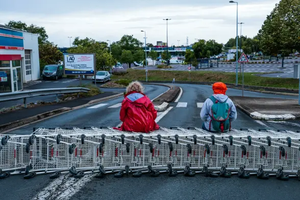 Deux manifestant·e·s sont assis·e·s sur une rangée de chariot de supermarché, au travers une route sans circulation.
L'un·e en imperméable rouge, et l'autre avec une capuche orange, un sweat gris et un sac à dos bleu/vert.
On peut apercevoir l'enseigne Intersport en arrière-plan.