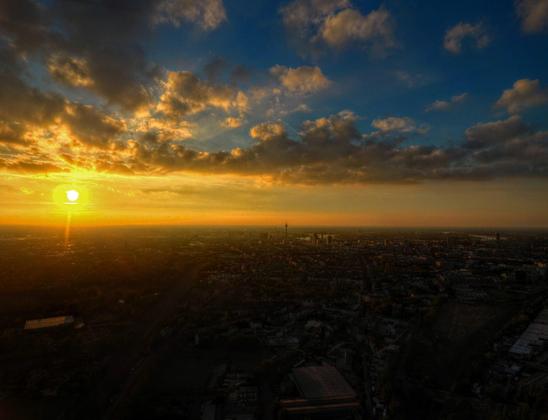 Foto: Luftbild eines Sonnenuntergang über eiem Großstadt-panomama. Im Vordergrund rechts Industriebrache/eheamaliges Bahn-Gelände, Bildmittig ein Kopfturm. Masten von Schrägseil-Betonbrücken. Sonne hinterleuchtet Wolken.
