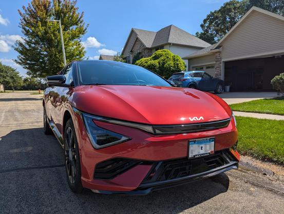 A shiny red Kia EV6 electric car parked on a suburban street in front of houses, with a tree and blue sky in the background.