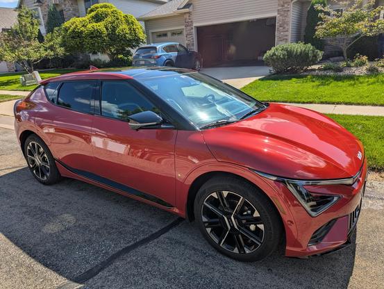 A red Kia EV6 electric car parked on a residential street, shown from a front-side angle with black wheels and tinted windows, in front of houses with green lawns and trees.