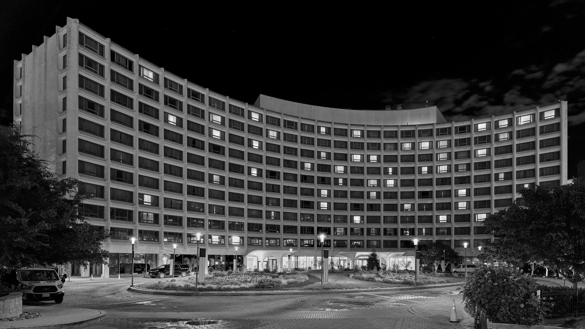 An eleven story midcentury modernist / brutalist hotel building. It has a curved facade and a plaza/driveway in front. It is night, and lights are visible in some of windows.