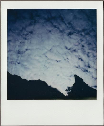 Polaroid looking up at blue sky covered in small white clouds, light from the sun behind the clouds illuminating a cliff edge at the bottom.