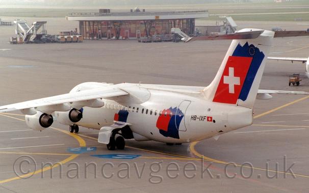 Rear side view of a high-winged, 4 engined jet airliner taxing from right to left.
The plane is mostly white, with large dark blue and small red splotches on the mid fuselage, over the main undercarriage, and a larger version on the rear fuselage, partially covering the rear cabin door.
An even larger version of the splotches adorn the tail, with the red splotch replaced by a Swiss flag, a red field with a large white cross in the middle., and the blue splotch behind it has a small circle of golden stars in the top right corner.
The black registration "HB-IXF" is on the rear fuselage, next to a small Swiss flag.
The foreground and most of the background is filled with grey concrete apron, with a circular terminal building in the background, surrounded by mobile air stairs.
There are large areas of grass lining a black runway in the distance, with trees vanishing into haze beyond that.