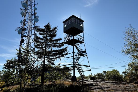 A tall metal fire lookout tower stands on a rocky hilltop surrounded by evergreen trees, with a small enclosed observation cabin perched at the top and accessed by a zigzagging staircase. Nearby, a telecommunications tower rises slightly higher, outfitted with multiple antennas and satellite dishes pointing in various directions. The terrain is rugged and sunlit, with exposed rock and sparse vegetation. The sky is clear and bright blue, suggesting a calm, sunny day.