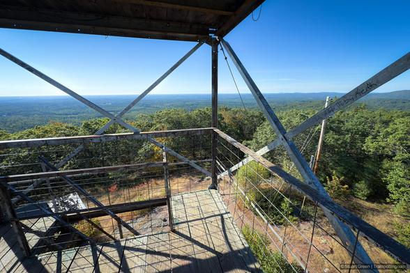 View from inside a fire lookout tower perched on a forested hilltop, showing its metal beam framework, wire fencing, and wooden floor and roof. The perspective looks outward through the open structure, revealing a sweeping landscape of densely wooded terrain stretching to the horizon under a clear blue sky. A utility pole with wires stands to the right, hinting at nearby infrastructure. The scene conveys a sense of elevated solitude and vigilance, with the tower’s rugged design emphasizing its purpose for wildfire monitoring and panoramic visibility.