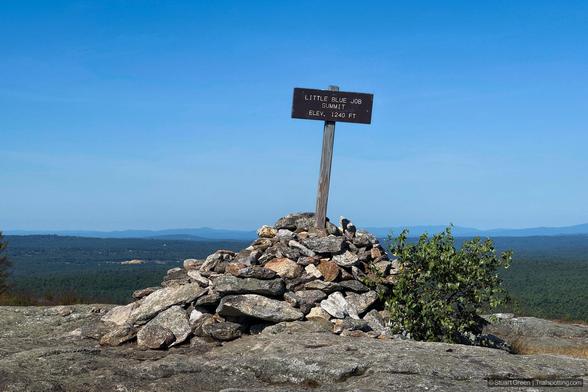 A summit marker stands atop a rugged pile of rocks at the peak of Little Blue Job Mountain. The marker is a weathered wooden sign mounted on a post, reading “LITTLE BLUE JOB SUMMIT ELEV. 1240 FT” in bold, carved lettering. The cairn beneath it consists of stacked stones, likely placed by hikers to commemorate their ascent. In the background, a sweeping view of forested hills stretches into the distance beneath a clear blue sky.