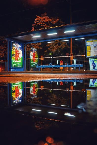 Das Bild zeigt eine nächtliche Szene an einer Bushaltestelle. Die Haltestelle ist modern gestaltet und besteht aus einer transparenten Glasstruktur mit blauen Sitzgelegenheiten. Sie wird von mehreren hellen Deckenleuchten beleuchtet, wodurch die Umgebung klar sichtbar ist. Auf den Glaswänden befinden sich verschiedene Plakate, darunter eine auffällig bunte Werbung für die neue Saison der Fußball-Bundesliga in der Sportschau. Vor der Haltestelle befindet sich eine große Pfütze, die das gesamte Bauwerk samt Beleuchtung und Plakaten spiegelbildlich reflektiert. Diese Spiegelung erzeugt eine symmetrische und visuell eindrucksvolle Wirkung. Im Hintergrund sind Bäume und Straßenlaternen zu erkennen, die die urbane Atmosphäre bei Nacht unterstreichen. Es sind keine Menschen oder Fahrzeuge im Bild zu sehen.