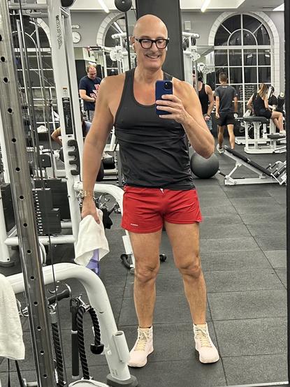 A person standing in a gym (me), taking a selfie. They are wearing a black tank top, red shorts, and light-colored athletic shoes. Various gym equipment and people working out are visible in the background.