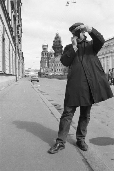 A standing person in pea coat taking a photograph of the photographer.  In the back is a Russian Orthodox church with scaffold on the cupolas.  It's a black and white photo.