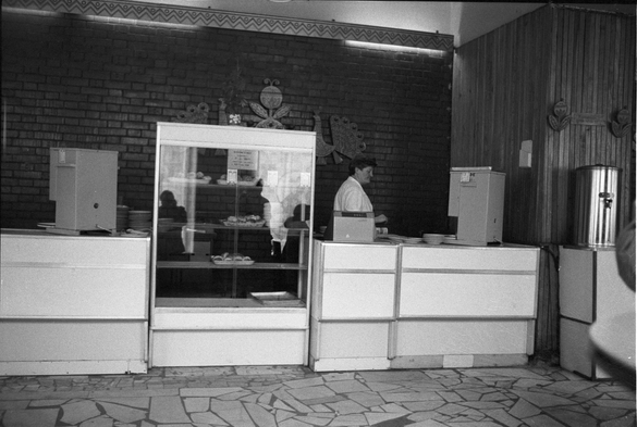 An interior of a cafe, stark, in black and white, with a solitary not happy looking worker behind the counter.  Few things in the case.