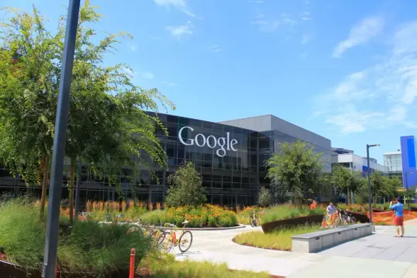 «Google»-Gebäude mit Glasfassade. Im Vordergrund ein Garten mit Bäumen, Gras und Blumen. Ein paar Leute und Fahrräder sind auf dem Weg zu sehen. Himmel ist blau und klar.