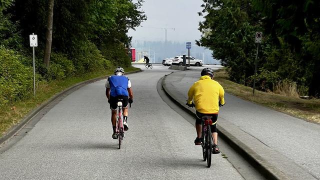 Two cyclists on a two-lane blacktop approaching a car parking area.