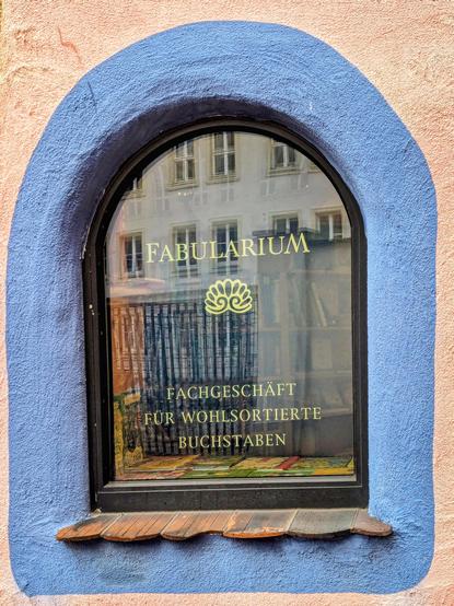 A close-up of a small, arched, black-framed window set into a textured wall painted in pale pink and blue. The blue paint forms a thick archway around the window. The reflection in the glass shows a building with large windows. The text on the window reads "FABULARIUM" in gold capital letters, followed by a gold circular logo resembling a tree or a seashell. Below that, in smaller gold letters, it says "FACHGESCHÄFT FÜR WOHLSORTIERTE BUCHSTABEN". Inside the window, you can see the spines of many books and other objects. The window's interior sill has books lying on it. The exterior sill consists of flat roof tiles.