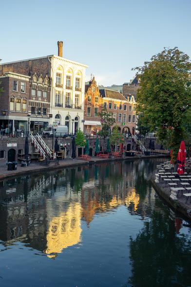The image captures a picturesque canal scene in an urban setting, showcasing reflections of architecture and nature. A row of historic buildings lines the far side of the canal, each with distinct architectural features; a prominent three-story white structure with ornate details stands out amidst its neighbors. The water appears calm, acting as a mirror to reflect the buildings, trees, and a clear sky. Bicycles are parked along the edge of the canal, with a few pedestrians visible. All the trees, with leaves in shades of green and orange-yellow, indicate an autumnal setting. Outdoor seating areas with red umbrellas suggest nearby cafes or restaurants. The scene suggests an urban space designed for leisure with clear skies casting a warm light, enhancing the vibrant colors and tranquility of the environment.
