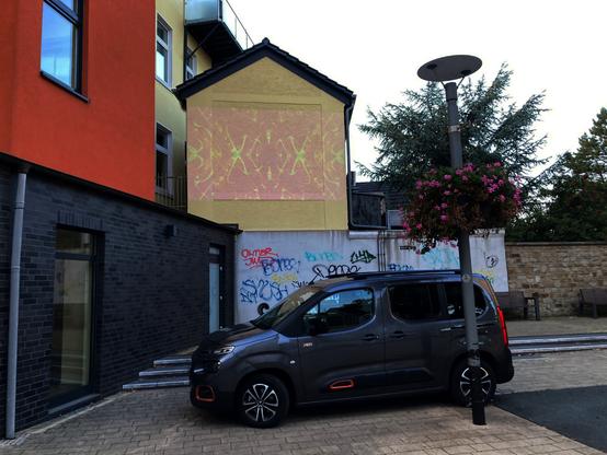 Farbfoto im Querformat. Auf die fensterlose Fassade eines kleinen Gebäudes ist ein Ljapunow-Diagram projiziert.