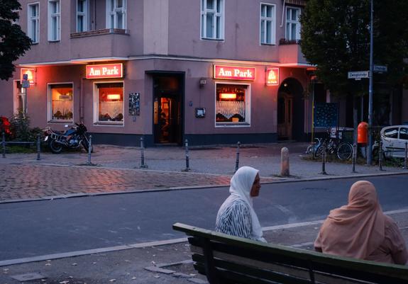 Eine Abendszene, die zwei Frauen zeigt, die auf einer Bank vor einem Gebäude mit beleuchteten Schildern mit der Aufschrift "Am Park" sitzen. Ein Motorrad ist in der Nähe geparkt, und die Straße ist mit Bäumen und Fahrrädern gesäumt. Die Atmosphäre ist ruhig und urban, mit sanftem Licht.