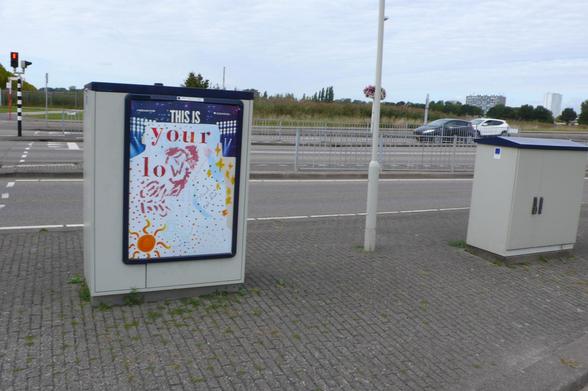 kale straat met twee straat kastjes voor autoweg met links een stoplicht, midden een paal en in de verte droog gekleurd gras. Op de een zit een bewerkt reclame bord, met boven nog wat reclame in donkere kleur met lichten schijnend naar beneden en witte letter: this is.
 recht zijkant wat geel. Het is aangepast in rode letters en links onder een oranje zon