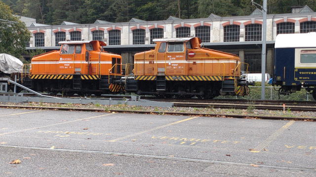 Zwei orangefarbene, dreiachsige Rangierdieselloks (mit unterschiedlichen Orangetönen bzw. unterschiedlichen Graden von Patina) stehen Heck an Heck vor einem Backstein-Industriegebäude.