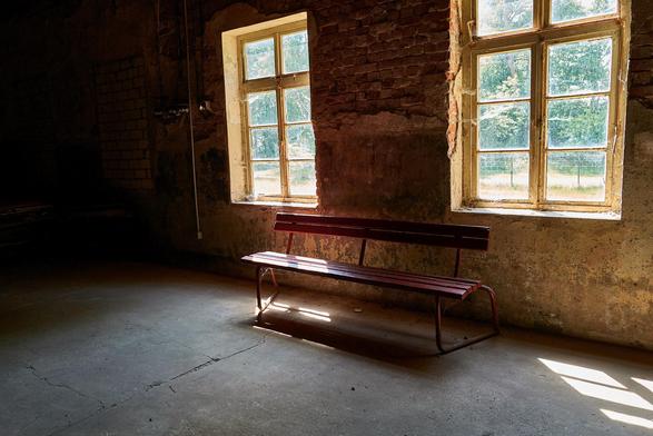 Farbaufnahme von einem kahlen Raum. Durch zwei Fenster fällt Sonnenschein hinein. Zwischen den Fenstern steht eine rote Bank. Ein Teil der Bank wird von der Sonne beschienen.
