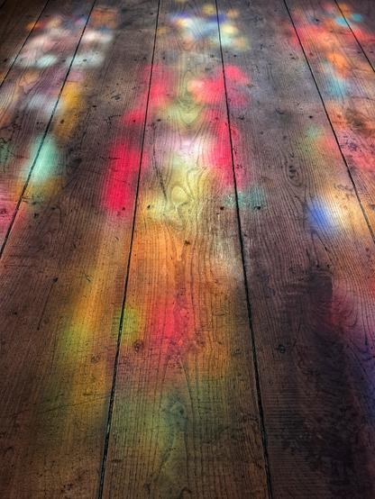 [pt] Reflexos coloridos de um vitral num chão de madeira

[en] Colorful reflections of a stained glass window on a wooden floor