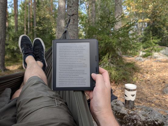 Kuva jaloistani riippumatossa, kädessä Kobon lukulaite ja viereisellä kivellä termosmuki. Taustalla metsää.