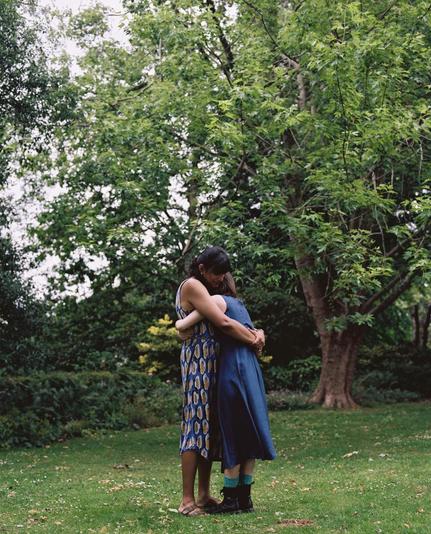 Two women hug each other in a garden with a large tree in the background.