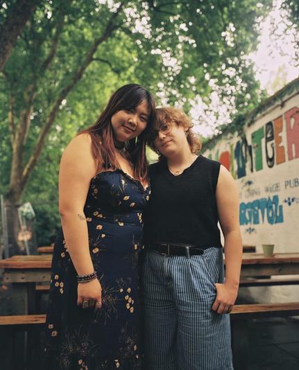 Two women stand close together outside the entrance to a pub, with pub tables and a tree behind them.
