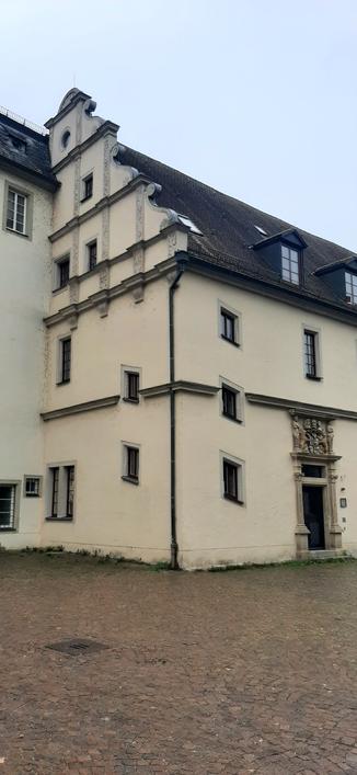 Das Bild zeigt einen Teil des Schlosses in Bad Mergentheim. Es ist ein mehrstöckiges Gebäude mit einer hellen Fassade und einem dunklen Dach. Die Fassade ist mit dekorativen Elementen verziert, wie z.B. Sprossenfenster und ein kunstvoll gestaltetes Portal mit einem Wappen darüber. Der Eingang ist mit einem kunstvollen Relief verziert. 

Das Gebäude hat mehrere Fenster, die in regelmäßigen Abständen angeordnet sind. Die Fenster rechts und links der Hausecke sind schräg angeordnet, was auf einen dahinterliegende Treppenraum hindeuten dürfte.

Text mit Unterstützung von @altbot
