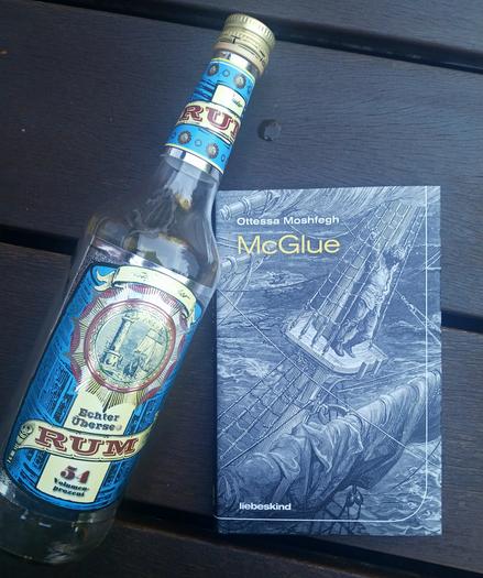 Foto des Buches, auf dessen Cover eine Schwarz-Weiß-Zeichnung zu sehen ist, auf der ein Mann bei sehr bewegter See oben auf dem Mast eines größeren Segelbootes steht und sich in den Wanten festhält. Er trägt Bart und längere Haare, seine Kleidung wirkt einfach und altmodisch. Das Buch liegt auf braunen Holzplanken, daneben eine fast leere Flasche »echter Übersee-Rum« auf dessen blau-gelben Etikett sich eine Plakette mit einer ähnlichen Schwarz-Weiß-Zeichnung findet: darauf ein Leuchtturm, ein Segelschiff und die Angabe »seit 1853«.