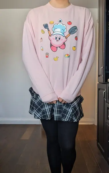I stand with black stockings and a partly covered black plaid skirt. I'm wearing a big comfy pink sweater. My hands are in front, holding the d bottom of the shirt. Pictures on the shirt is Kirby with a chef had, a flipper, frying pan, and is encircled with food