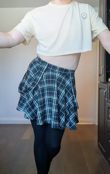 Black stockings and black plaid skirt, I rest in one leg and have my torso bend. I'm wearing a beige-ish, loose cropped T-shirt with a little smiley face on it