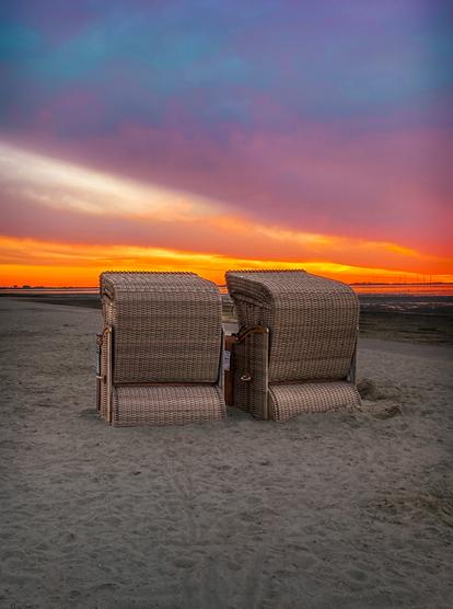 Zwei braune Strandkörbe stehen mit dem Rücken zum Betrachter schräg am Sandstrand. Es ist Ebbe an der Nordsee, man sieht den Schlick rechts von ihnen. Der Himmel ist stark bewölkt, die Wolken werden am Horizont von der untergehenden Sonne in ein leuchtendes Orange getaucht. Ein fast weißer Streifen markiert den Übergang in ein blau-lilafarbenes Farbspktrum.