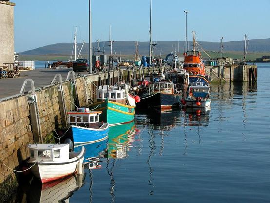 The pier at Stromness in Orkney. The image shows the pier entering the lower left of the frame and curving around to the right. Numerous boats are moored to the pier, mostly facing the viewer. One of them is a lifeboat. In the background are two levels of land, a low island and a rather higher one beyond it. The sky is blue.