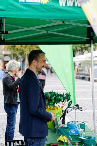 Fotografie von zwei Personen. Im Vordergrund eine männliche Person im blauen Jacket unter dem Pavillon der Grünen. Vor ihm liegt auf einer Art Theke Wahlkampfmaterial. Im Hintergrund stehen grüne Windräder in einem gelben Eimer. Im Hintergrund sieht man eine weibliche Person im Gespräch.