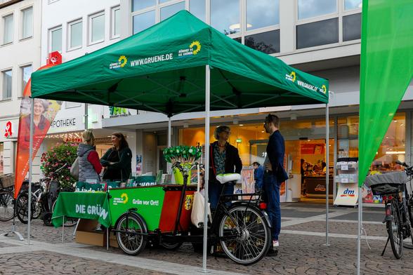 Fotografie eines Infostands der Grünen in Brühl vor einer DM-Filiale in der brühler Innenstadt. Man sieht das grüne Lastenfahrrad, das als Tisch dient, den grünen Pavillon mit dem Logo von Bündnis90/Die Grünen und einige grüne Wiindräder auf dem Lastenfahrrad. Links und rechts des Pavillons stehen Beachflags. Die linke Beachflag zeigt ein Portait von Simone Holderried. Unter dem Pavillon stehen vier Personen paarweise im Gespräch.