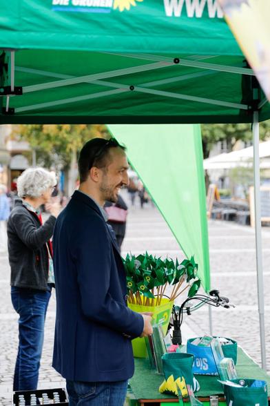 Fotografie von zwei Personen. Im Vordergrund eine männliche Person im blauen Jacket unter dem Pavillon der Grünen. Vor ihm liegt auf einer Art Theke Wahlkampfmaterial. Im Hintergrund stehen grüne Windräder in einem gelben Eimer. Im Hintergrund sieht man eine weibliche Person im Gespräch.