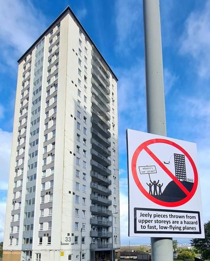 A sign beside block of high rise flats, featuring a high rise flat with kida protesting outside it. Underneath are the words Jeely piecea thrown from upper storeys are a hazard to fast, low-flying planes (which is a lyric from the Jeely Piece Song).