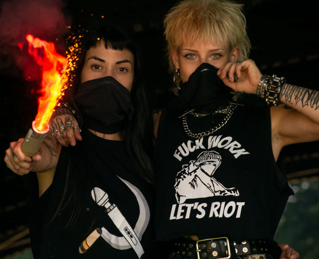 An image of two people wearing black shirts. one of the shirts has a hammer and sickle with the hammer being a vibrator, the other says “fuck work, let's riot”