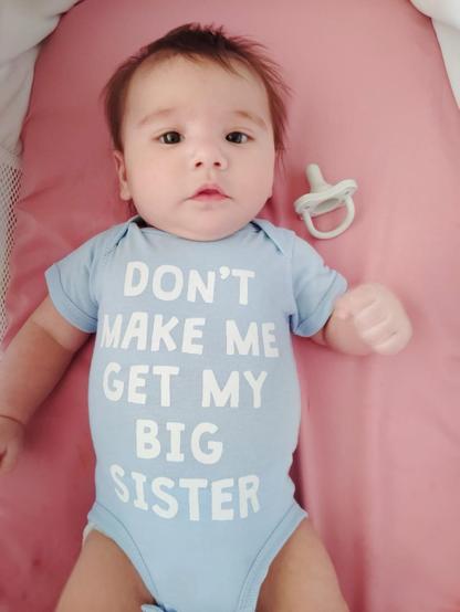 A light skinned baby with dark hair who is about 2 months old in a blue onesie that says “Don’t make me get my big sister.” There is a binky beside him in the bassinet.