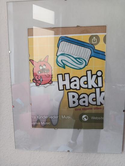 Eingerahmter Ausschnitt eines Posters mit HackiBacki, ein rotes Karies-Monster mit Spizhacke, das auf einem Zahn sitzt und von einer Zahnbürste bedroht wird. Es handelt sich dabei wohl um ein Zahnputz-Kinderlied. Das zweite "i" ist vom Rahmen verdeckt.