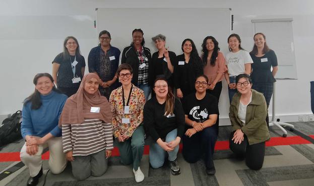 “Smiling group of 14 R-Ladies at R Dev Day, posing together in a meeting room with a whiteboard and chairs in the background.”