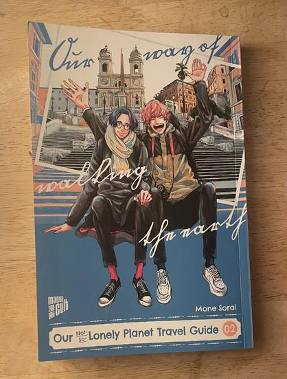 Photo of the manga our not so lonely planet travel guide volume 2 by Mone Sorai, German edition by Manga Cult. The cover shows two men sitting on a Staircase in Rome. One has black hair and wears glasses and looks rather reserved but smiles at the other. The otherha swabs shirt pink hair and he is waving and smiling happily .
