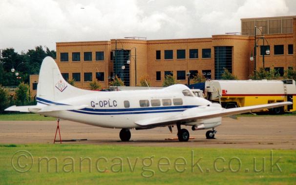 Side view of a small, white, twin propellor-engined airliner parked facing to the right and slightly away from the camera.
The plane is mostly white, with a double blue stripe running along the body, and the blue registration "G-OPLC" on the upper rear fuselage.
The cockpit is contained in a bubble higher than the passenger cabin, as can be seen with the difference between the cockpit and cabin windows.
A triangular red frame is propping up the tail, resting on the ground.
A white and red fuel tanker is parked in the background on the right of the frame, in front of a sandy-brown brick building with large, black windows.
Cloudy grey sky fills the rest of the frame.