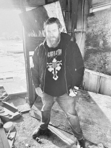 Josh Henderson of The Final Witness, photographed in black and white. Standing in a weathered, half-collapsed building, he wears a Tribulation shirt and jacket, looking directly at the camera. Henderson is the driving force behind The Final Witness, handling the band's creative and musical direction.