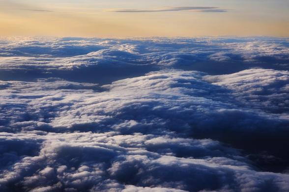 A wide aerial view of a dense, textured layer of clouds from above. The clouds are lit from the top by the golden light of the setting sun, casting blue and purple shadows in the valleys between the puffy white clouds. The horizon is a light orange and pink, with a few faint clouds visible above the main cloud layer.