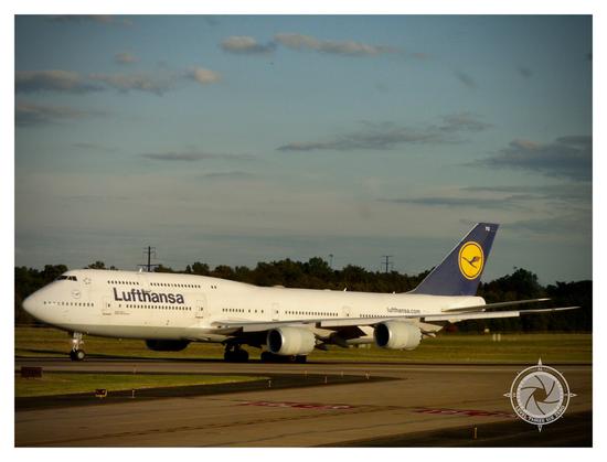 An almost side view of a Lufthansa Boeing 747-8 rolling down the runway just prior to takeoff. The out of frame setting sun casts a golden light across the white fuselage. The sky is blue with a few cumulus clouds.