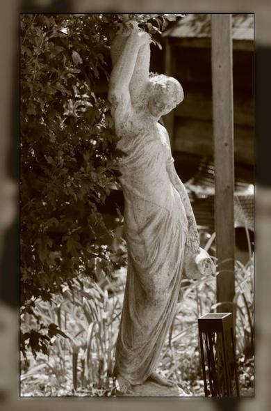 Garden ornament, faux Greek statuary of a woman in a flowing wrap, with a water jar on her shoulder. Beside the statue is a wooden post to the right, on the left is a large bush. In the background is a garden shed.